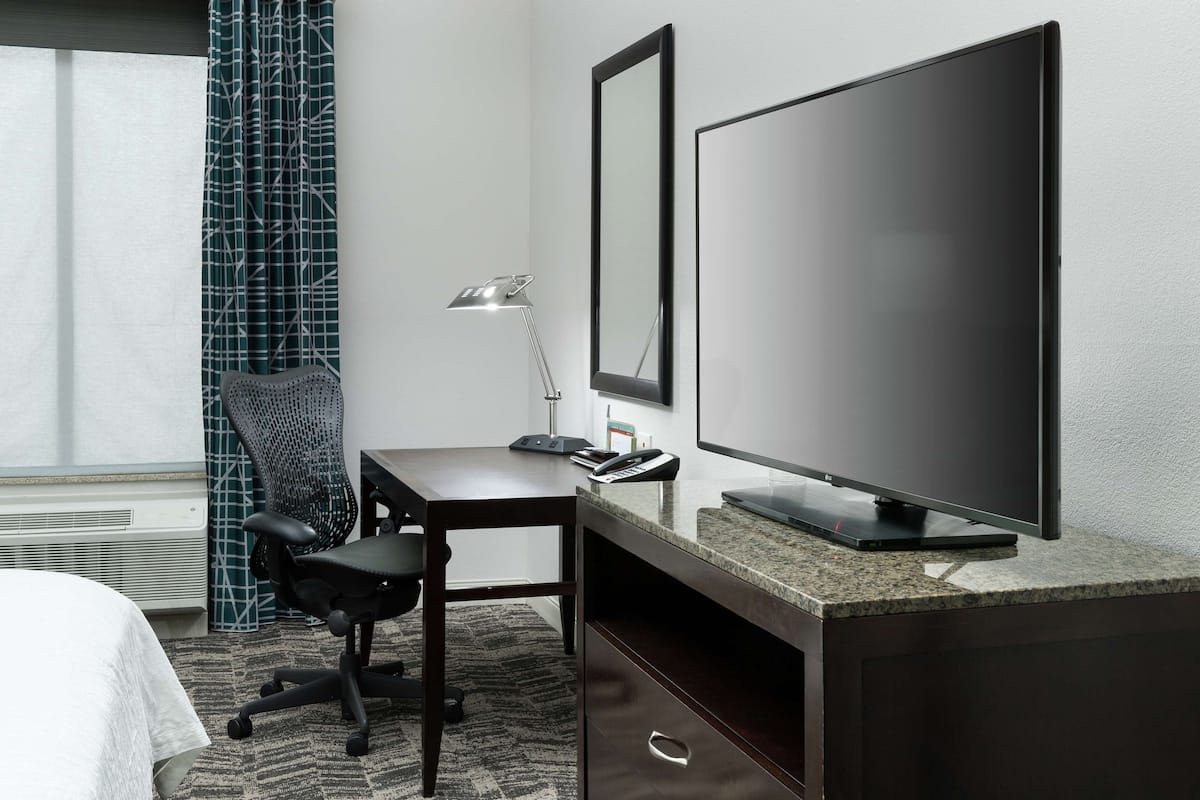 A hotel room with a bed, a desk with a computer, a TV, a lamp, and a chair.