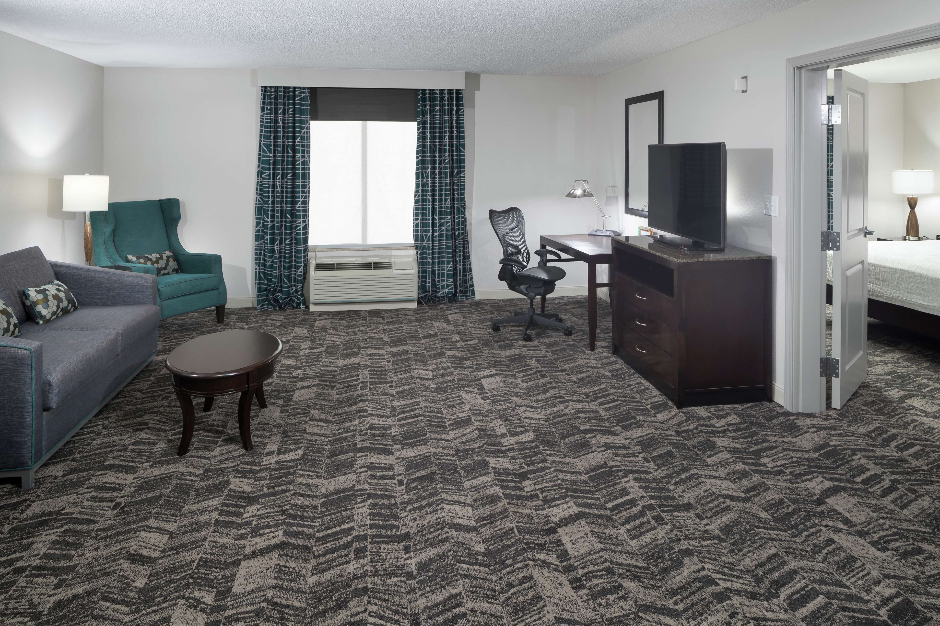 A hotel room with a grey sofa, teal armchair, and a desk with a TV.