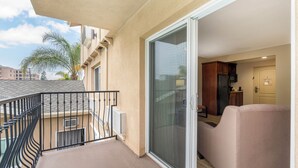 Un balcon doté d’une rambarde noire, une porte-fenêtre coulissante donnant sur un salon avec un canapé beige, et une vue sur des palmiers et des bâtiments en arrière-plan.
