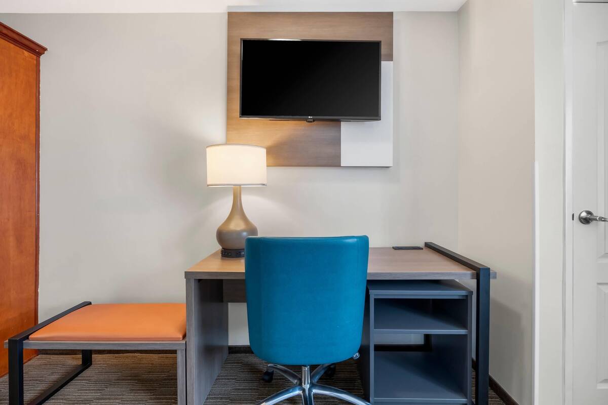 Een hotelkamer met een bureau, een blauwe stoel, een aan de muur bevestigde televisie en een lamp.