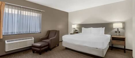 A hotel room with a large bed, two bedside tables with lamps, a chair, a window with curtains, and a wall-mounted heater.