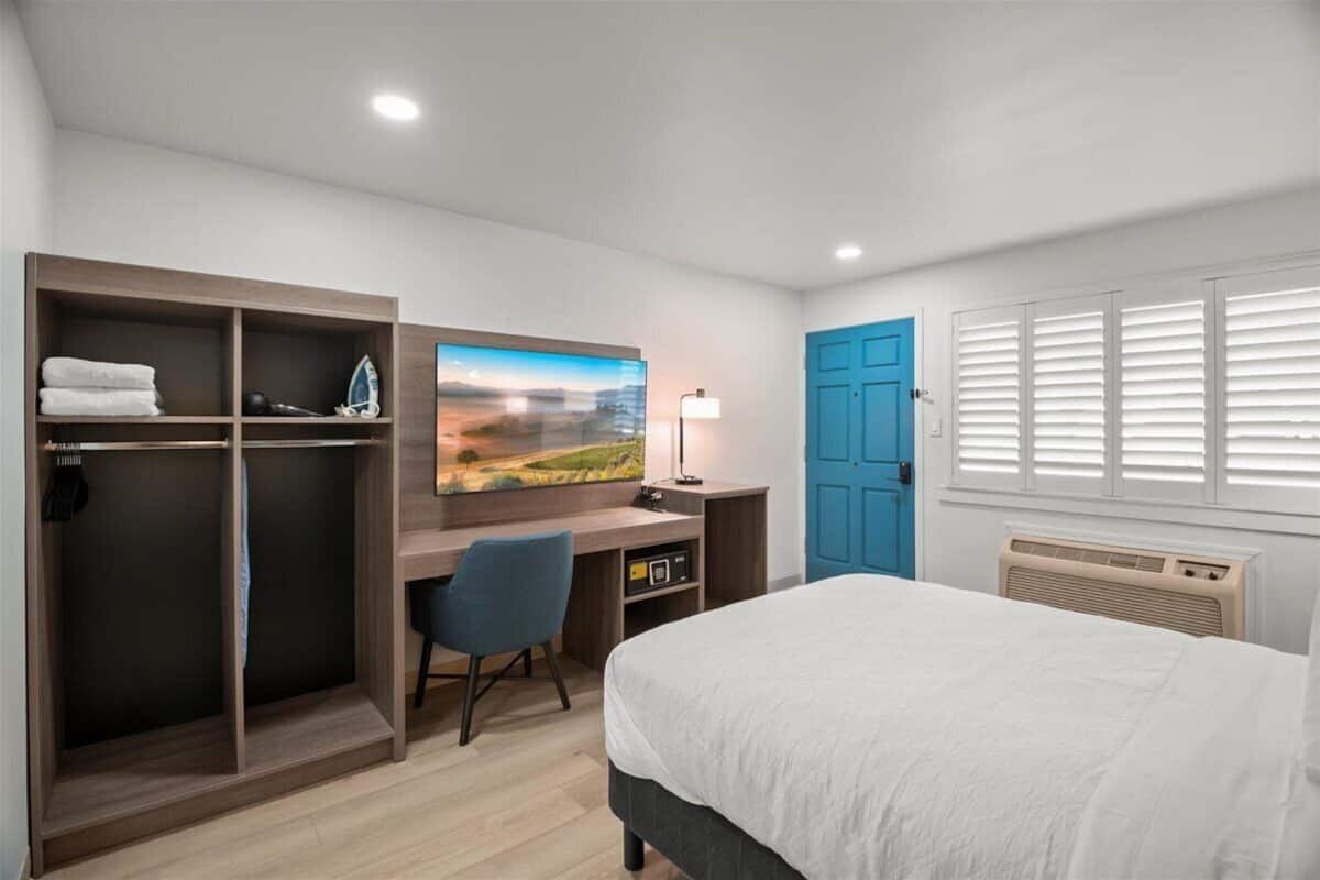 A bedroom with a bed, a desk with a chair, a wardrobe, a painting, a blue door, and a window with blinds.