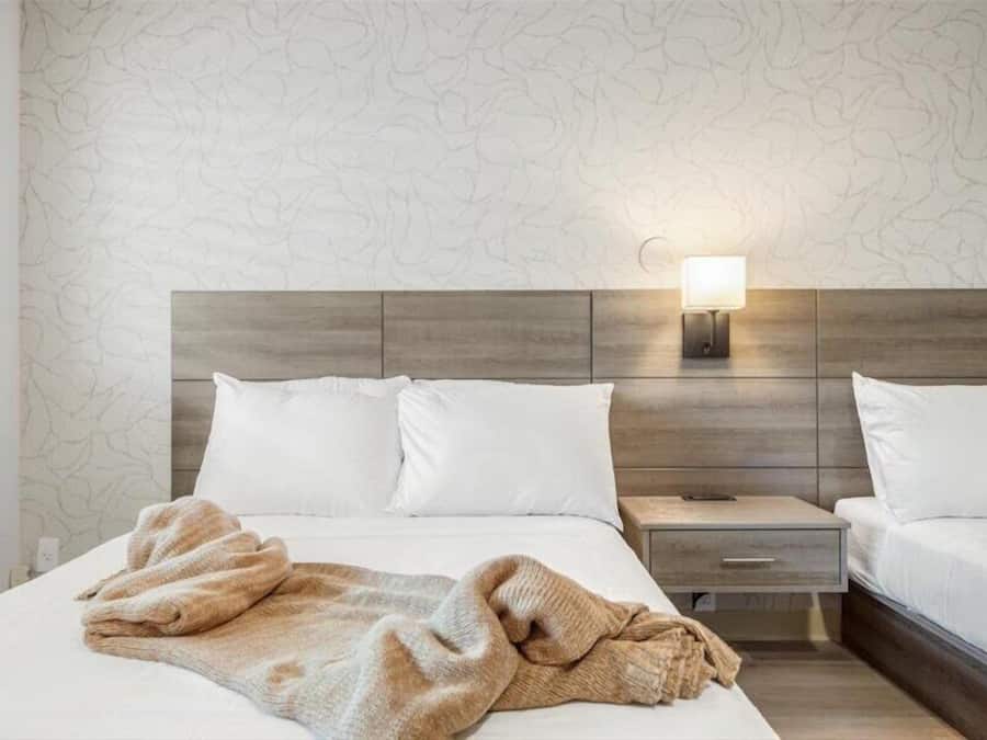 A hotel room with a bed, two pillows, a blanket, a nightstand, and a wall-mounted lamp.