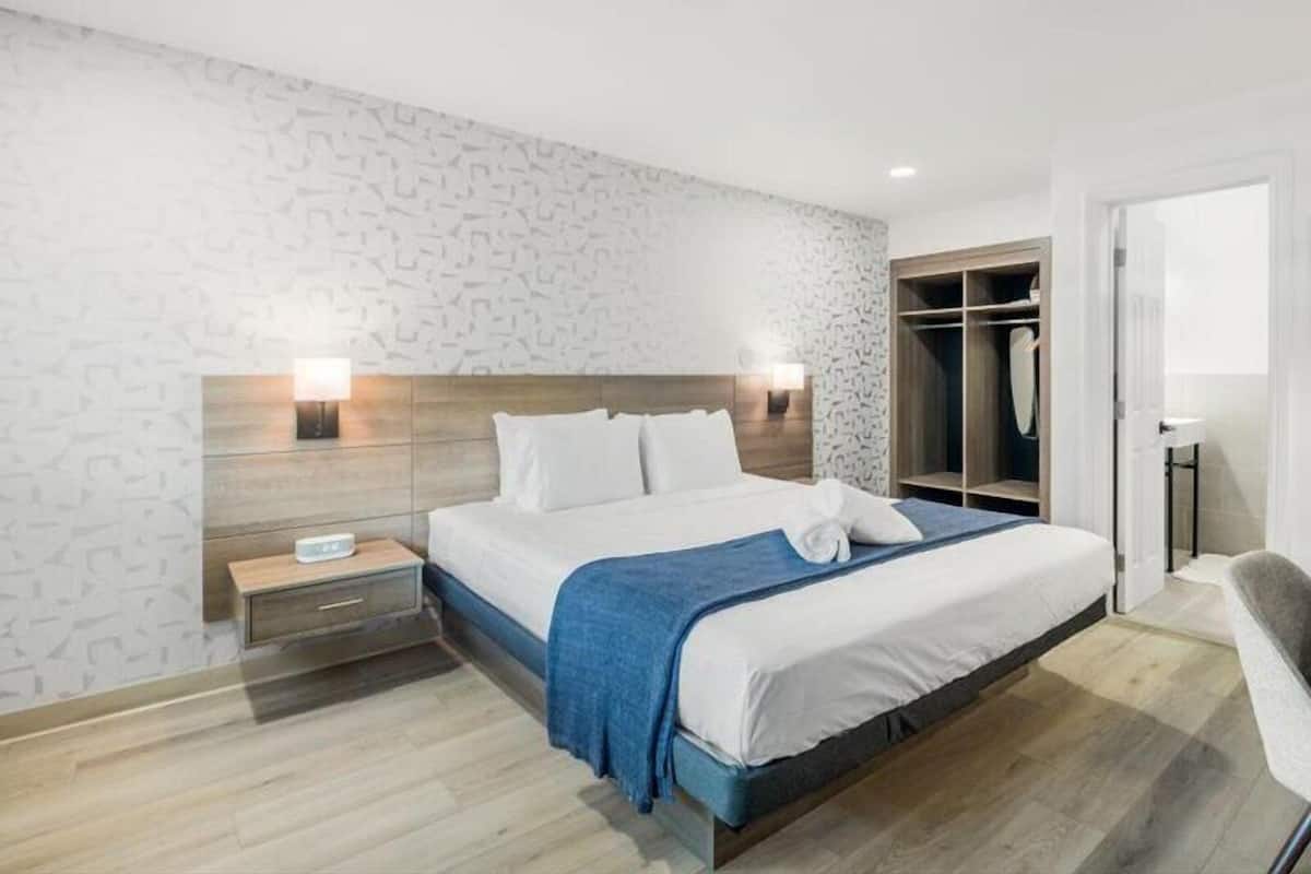 A modern hotel room with a large bed, bedside lamps, a wooden headboard, a walk-in closet, and a bathroom with a toilet and sink.