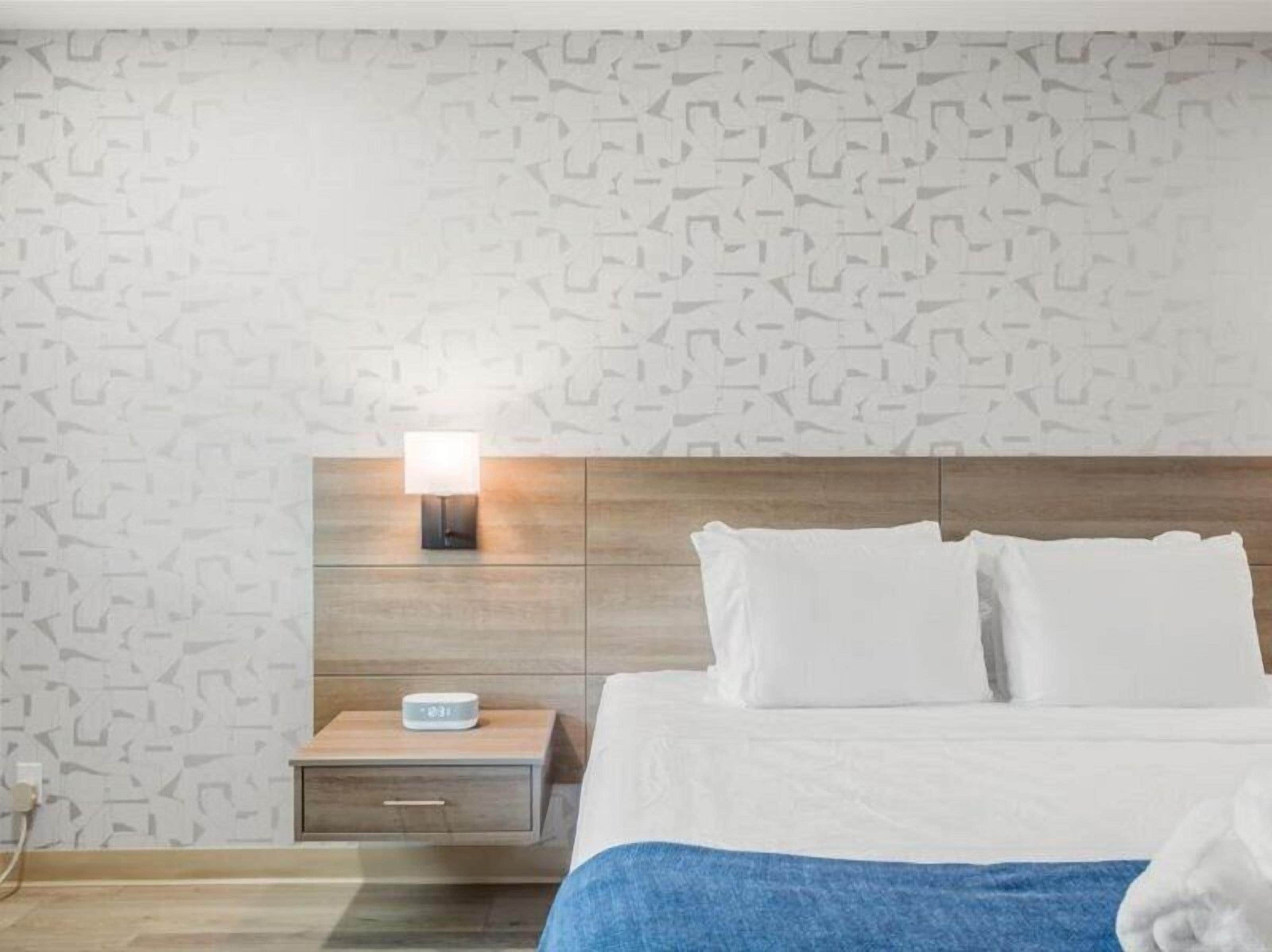 A hotel room with a patterned wall, a wooden headboard, a bedside table with a lamp, a digital clock, and a neatly made bed with white pillows.