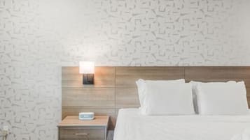 A hotel room with a patterned wall, a wooden headboard, a bedside table with a lamp, a digital clock, and a neatly made bed with white pillows.