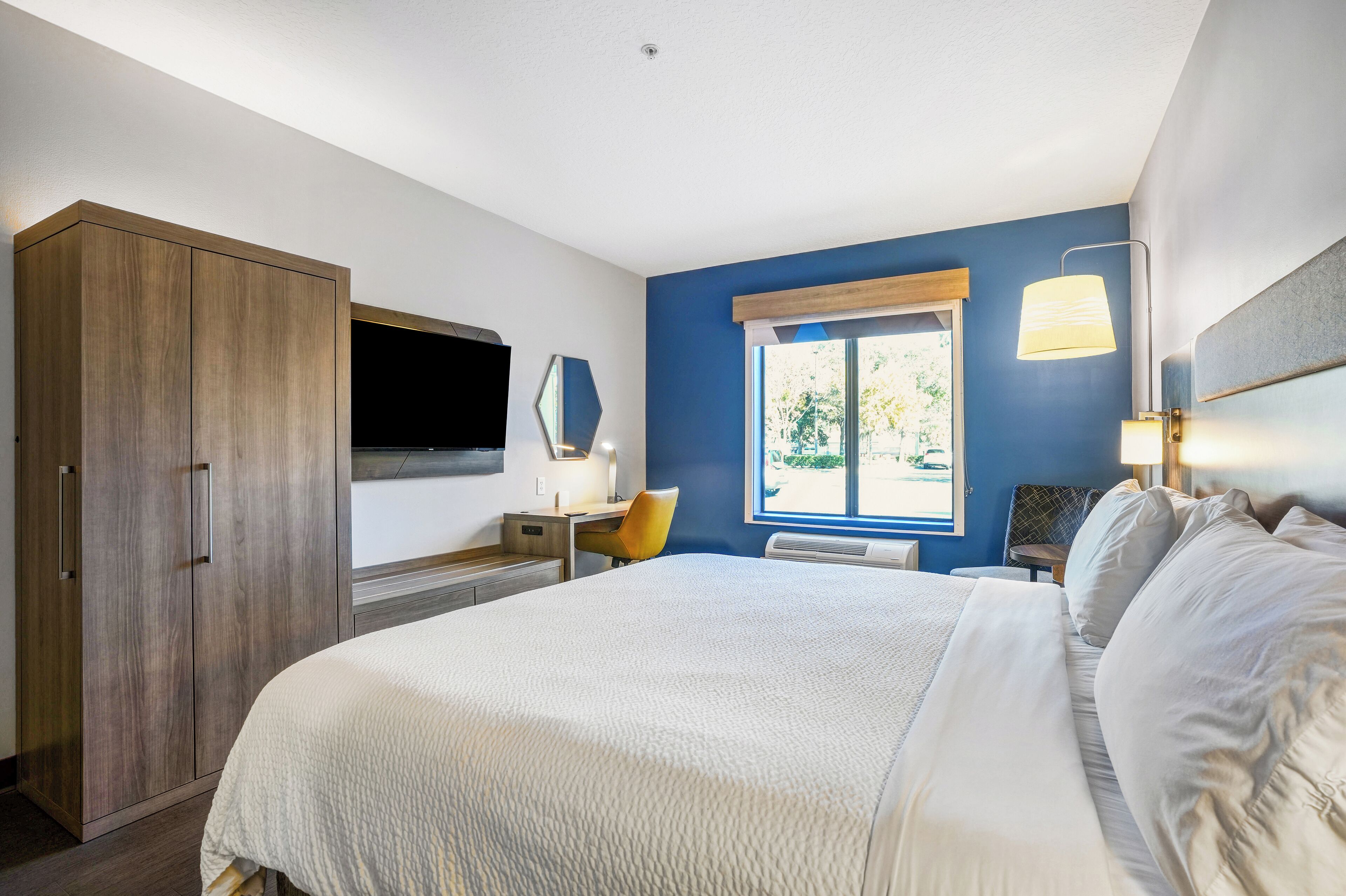 A bedroom with a large bed, a desk, a chair, a wardrobe, and a TV mounted on the wall.