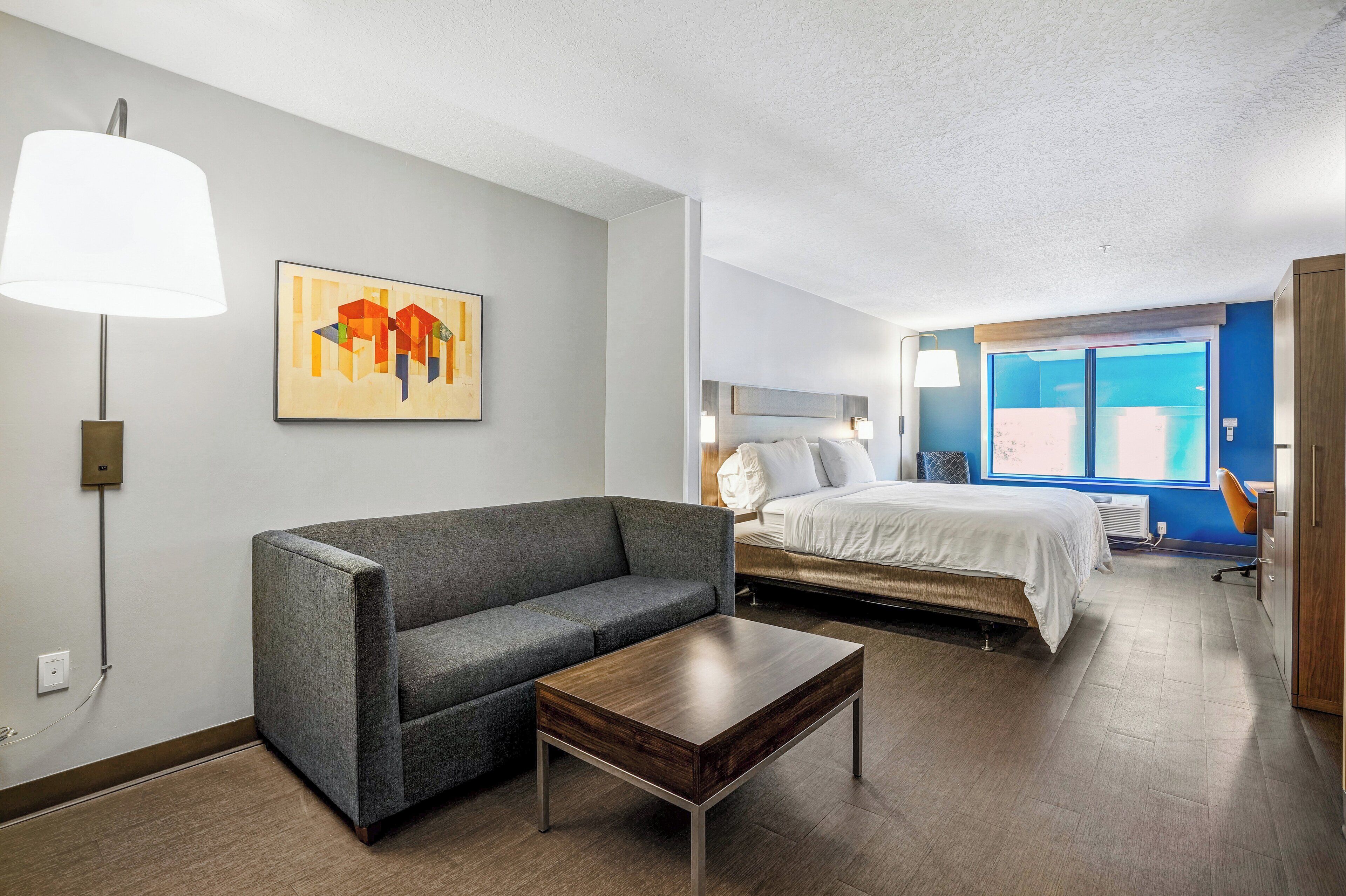 A modern hotel room with a grey sofa, a wooden coffee table, a bed with white bedding, a painting on the wall, and a large window with a view.