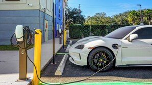 Un coche eléctrico blanco cargándose en una estación, con un edificio al fondo.