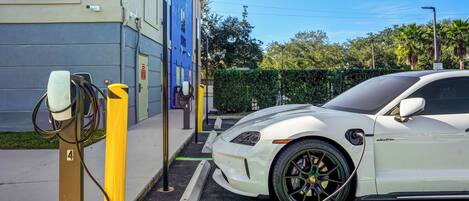 Une voiture électrique blanche est en train de se recharger à une borne, avec un bâtiment visible en arrière-plan.
