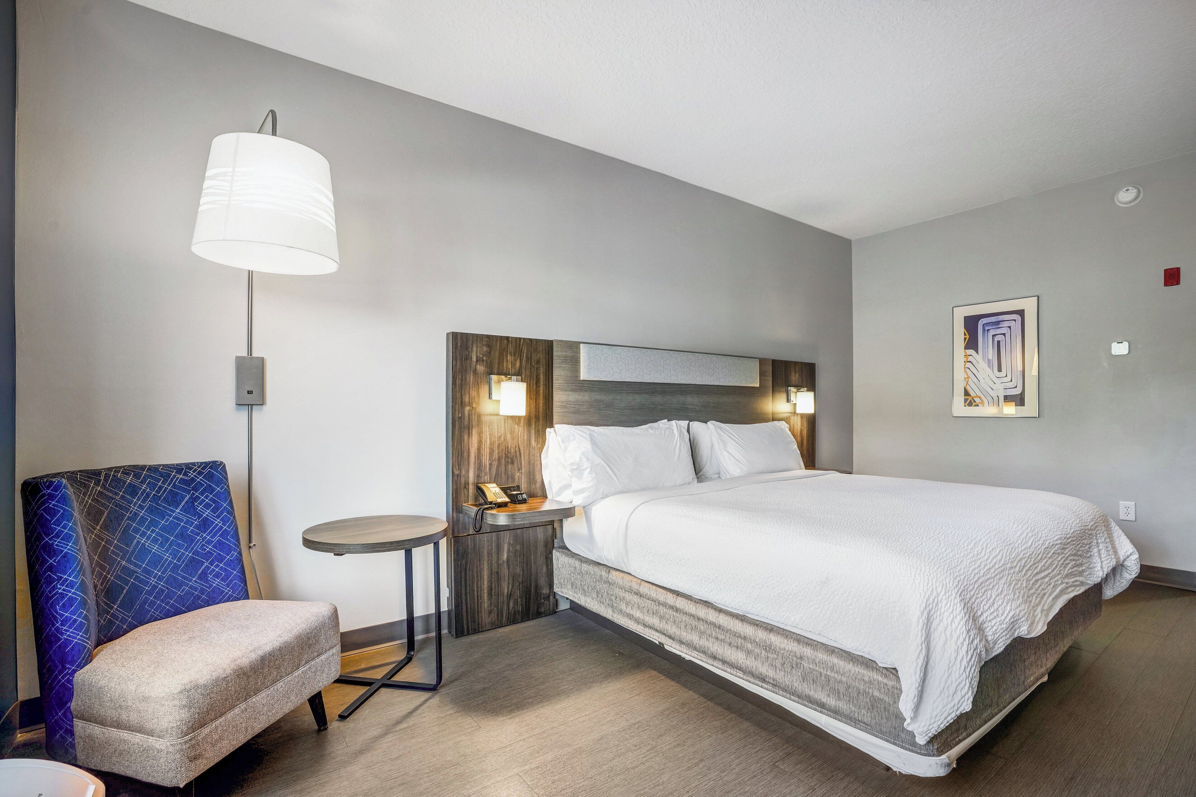 A hotel room with a bed, a chair, a bedside table, a lamp, and a wall-mounted artwork.