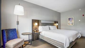 Quarto de hotel com uma cama, uma cadeira, mesa de cabeceira, abajur e uma obra de arte na parede.