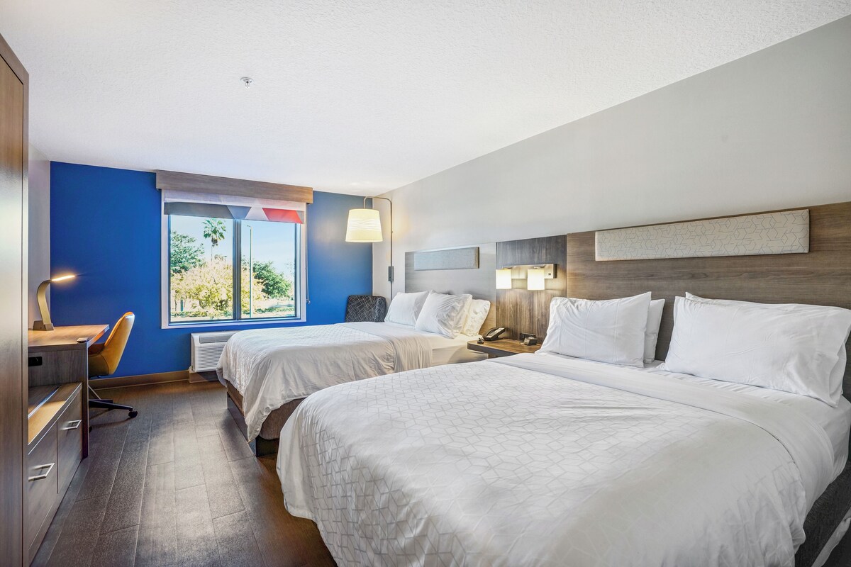 A hotel room with two beds, a desk, and a chair. There is a window with a view of trees and a lamp on the desk.