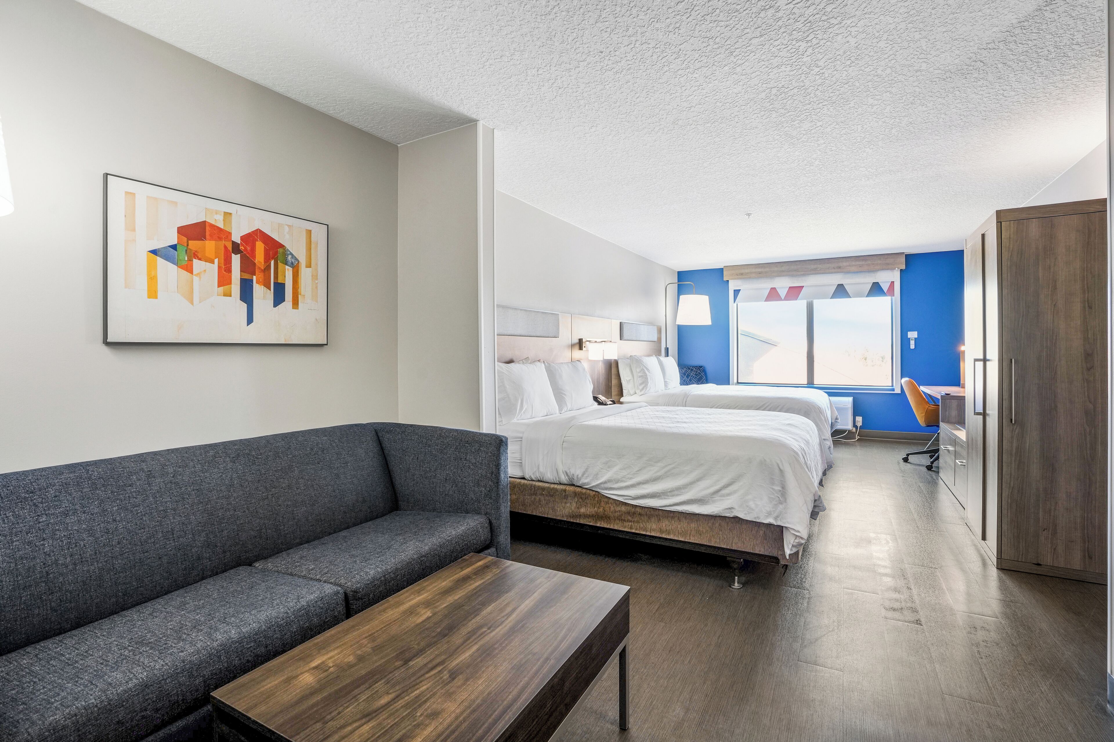 Una habitación de hotel moderna con un sofá gris, una mesa de madera, una cama doble con ropa blanca y una pared de acento azul, además de una ventana con cortinas.