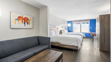 Una habitación de hotel moderna con un sofá gris, una mesa de madera, una cama doble con ropa blanca y una pared de acento azul, además de una ventana con cortinas.