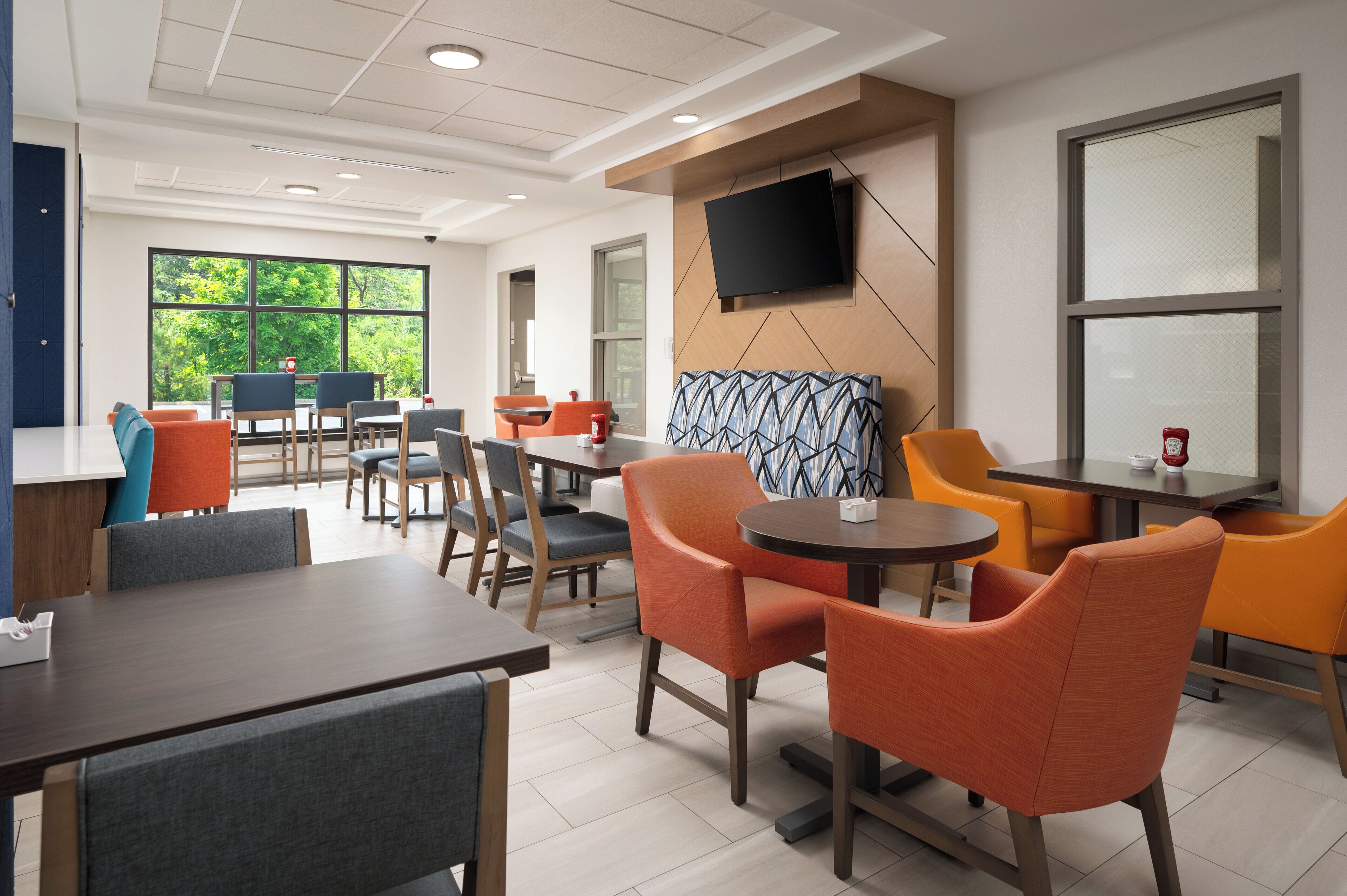 A modern dining area with multiple tables and chairs, a flat-screen TV mounted on the wall, and large windows offering a view of greenery outside.