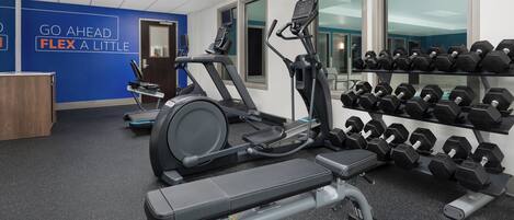 A gym with treadmills, an elliptical, and a bench press. There are dumbbells on a rack and a motivational quote on the wall.