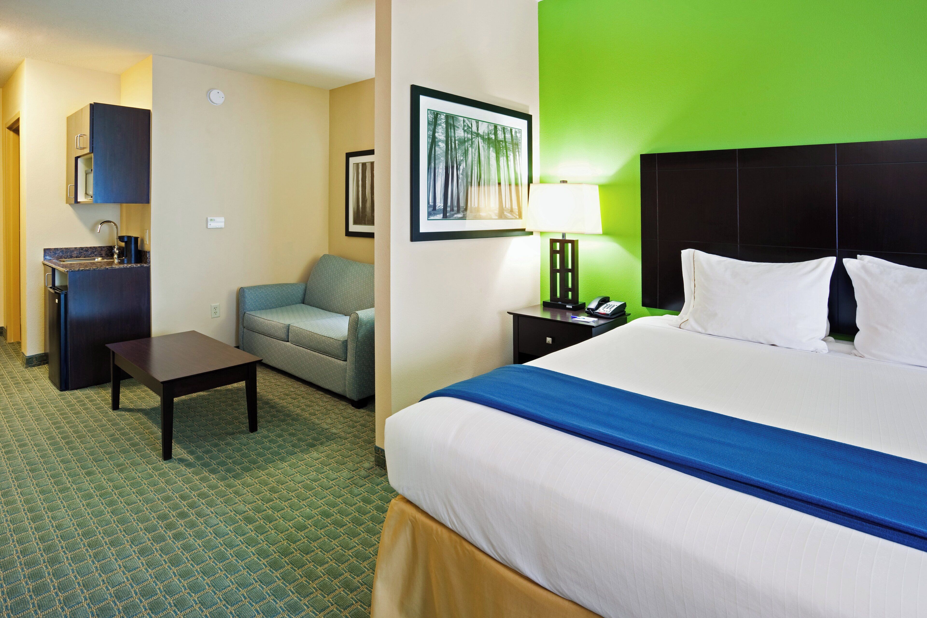 A hotel room with a bed, a desk, a chair, a sofa, a nightstand, a lamp, and a framed picture on the wall.