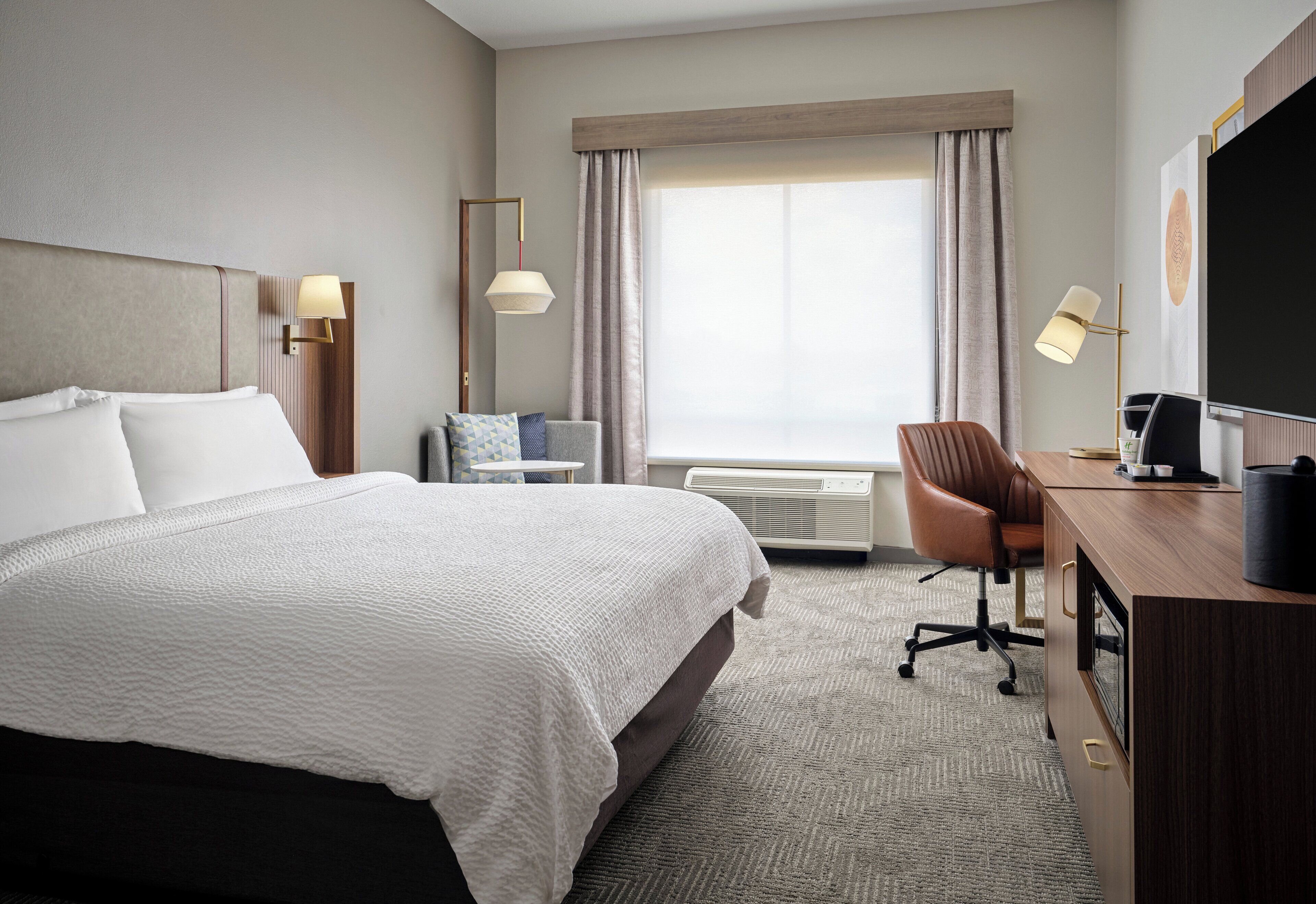 A hotel room with a large bed, a desk with a computer, a chair, a TV, and a window with curtains.