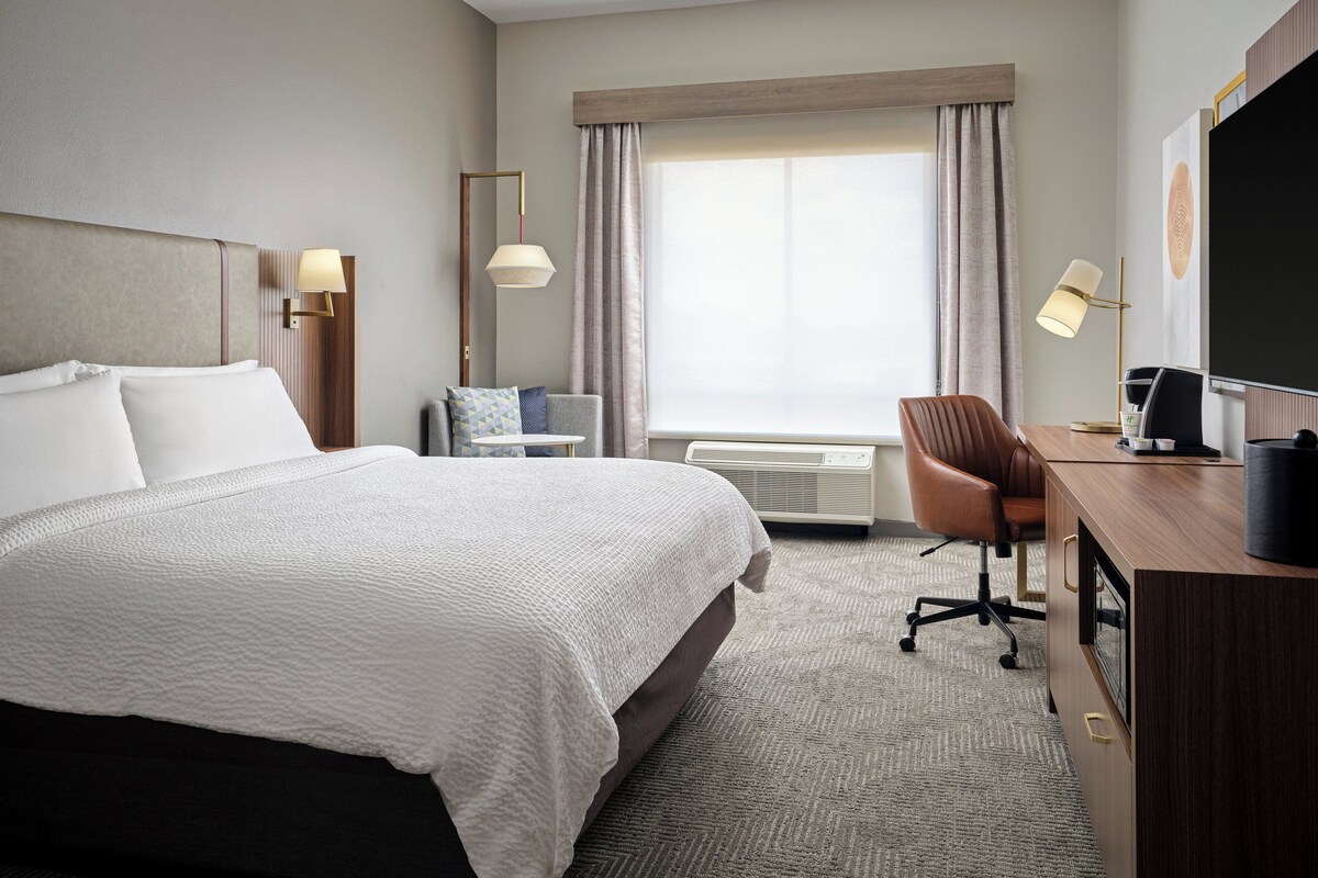 A hotel room with a large bed, a desk with a computer, a chair, a TV, and a window with curtains.
