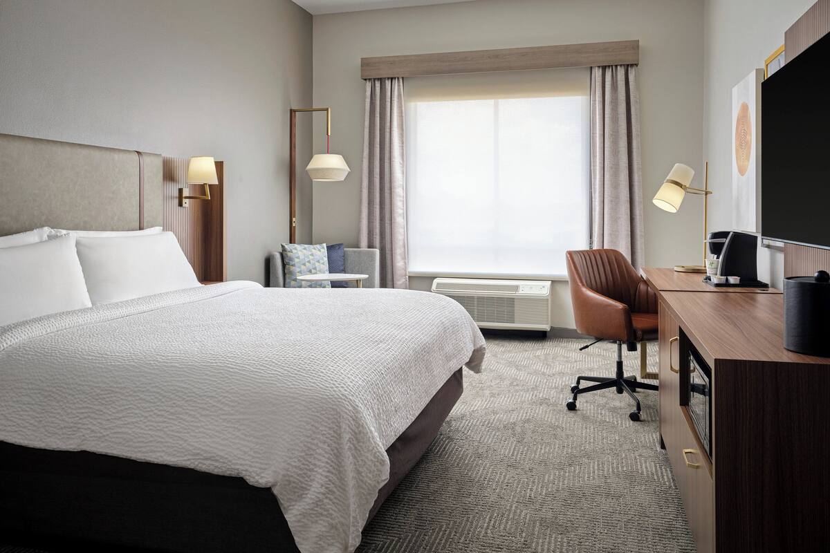 A hotel room with a large bed, a desk with a computer, a chair, a TV, and a window with curtains.