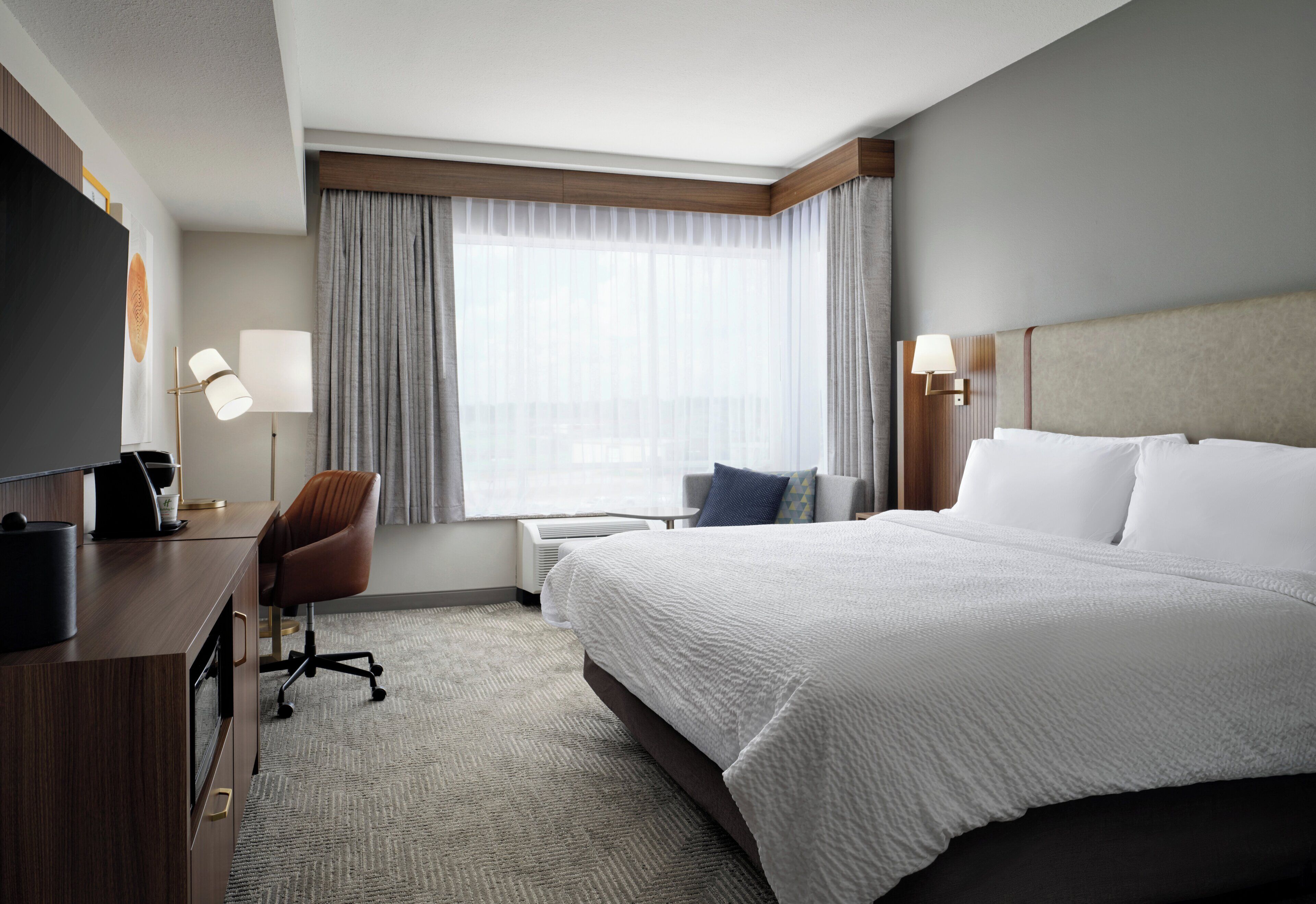 A hotel room with a large bed, a desk with a chair, a flat-screen TV, and a window with curtains.