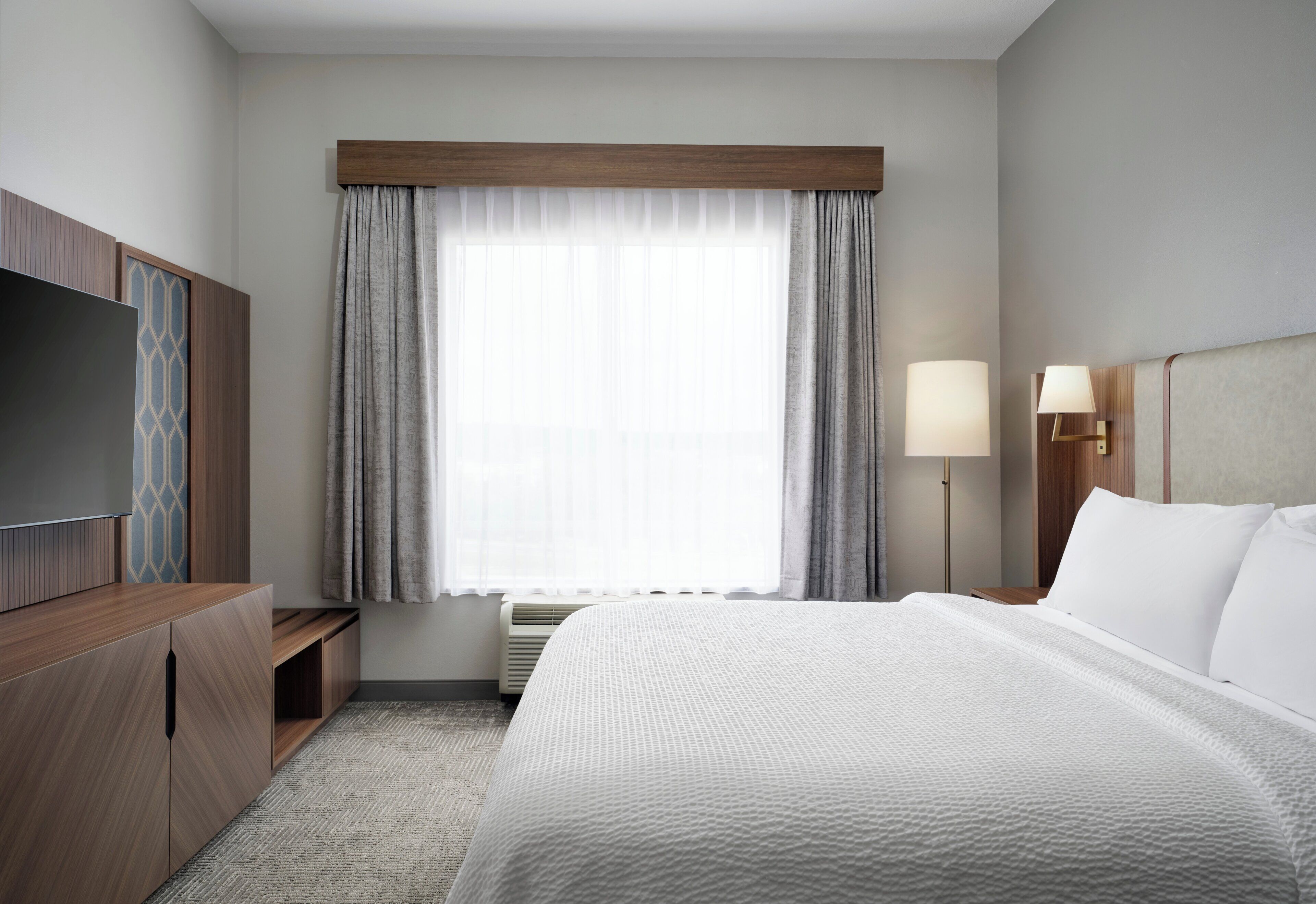 Ein Hotelzimmer mit einem großen Bett, einem Fernseher auf einer Holzkommode, einem Schreibtisch mit Lampe und einem mit Vorhängen versehenen Fenster.