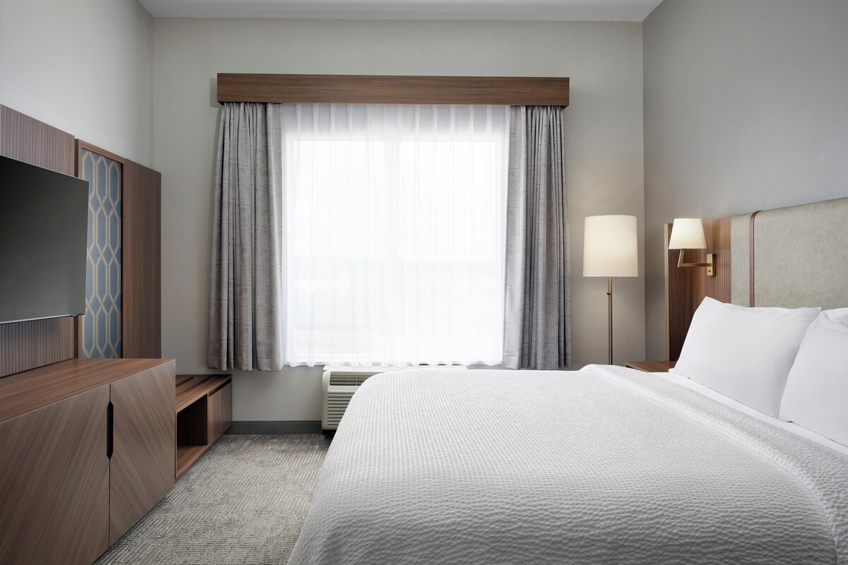 A hotel room with a large bed, a TV on a wooden cabinet, a desk with a lamp, and a window with curtains.