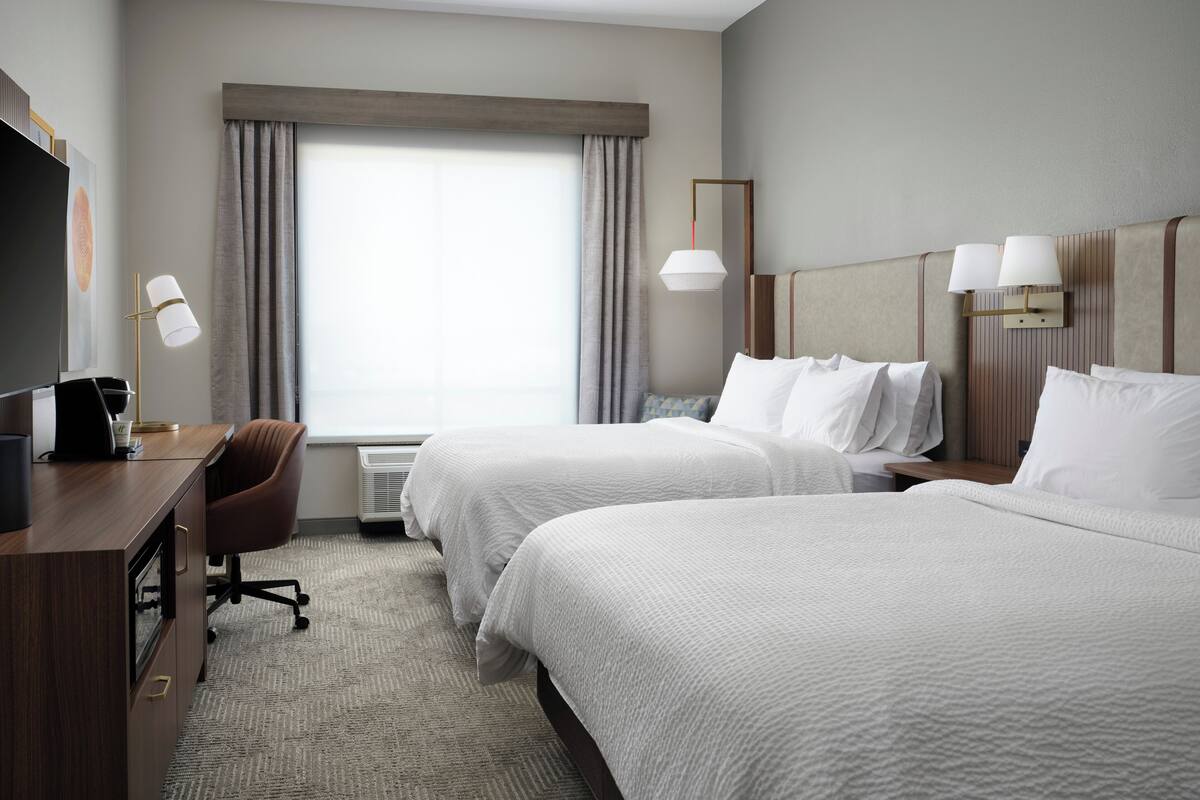 A hotel room with two beds, a desk with a chair, a television, and a window with curtains.
