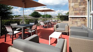 En udendørs terrasse med borde, stole og parasoller, omgivet af stenvægge og et metalhegn.