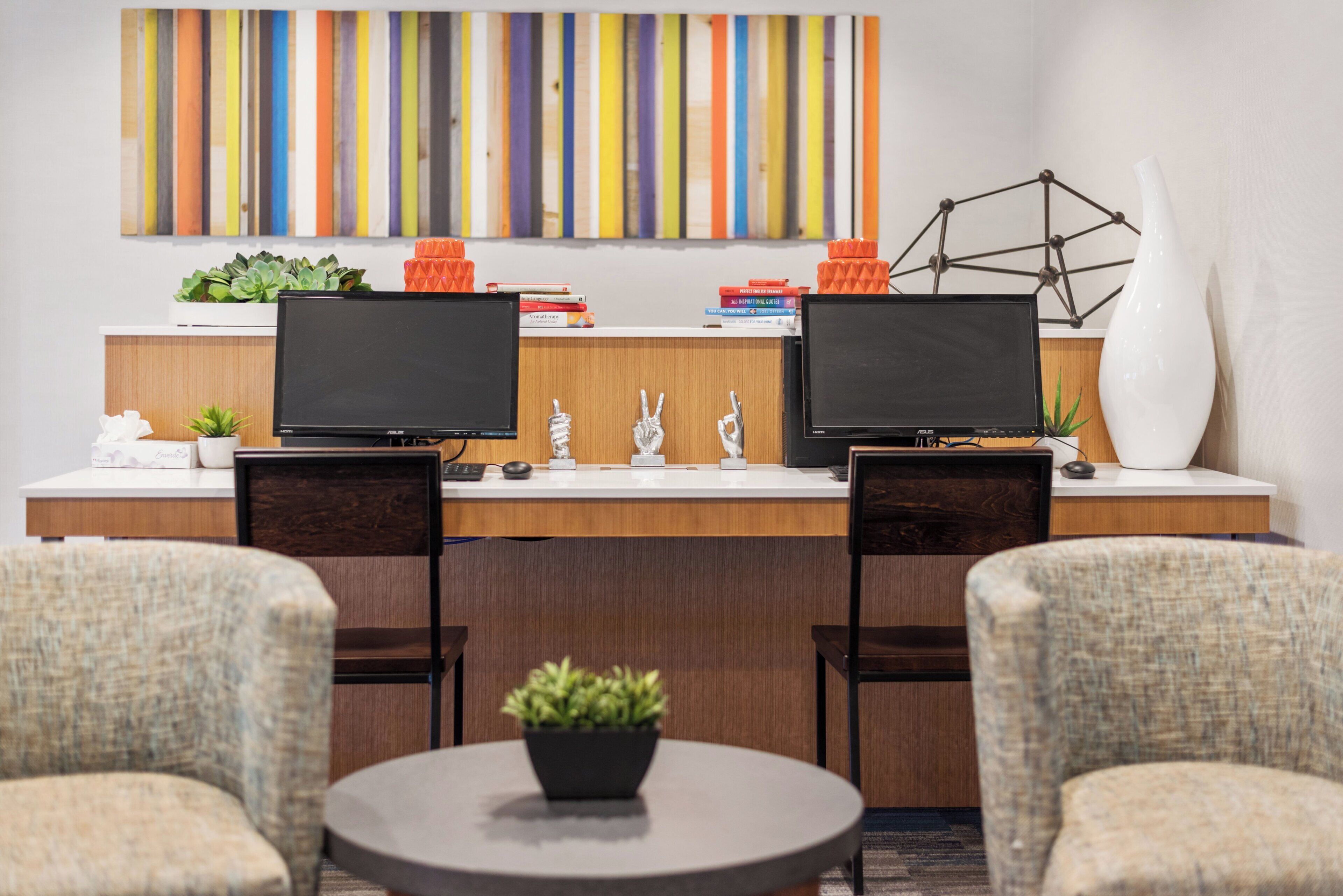 A modern office space with two monitors, a desk, two chairs, a small table with a plant, and a colorful abstract painting on the wall.