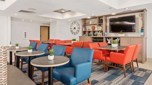 A hotel lobby with round tables, orange chairs, a large clock, a TV, and a bookshelf.
