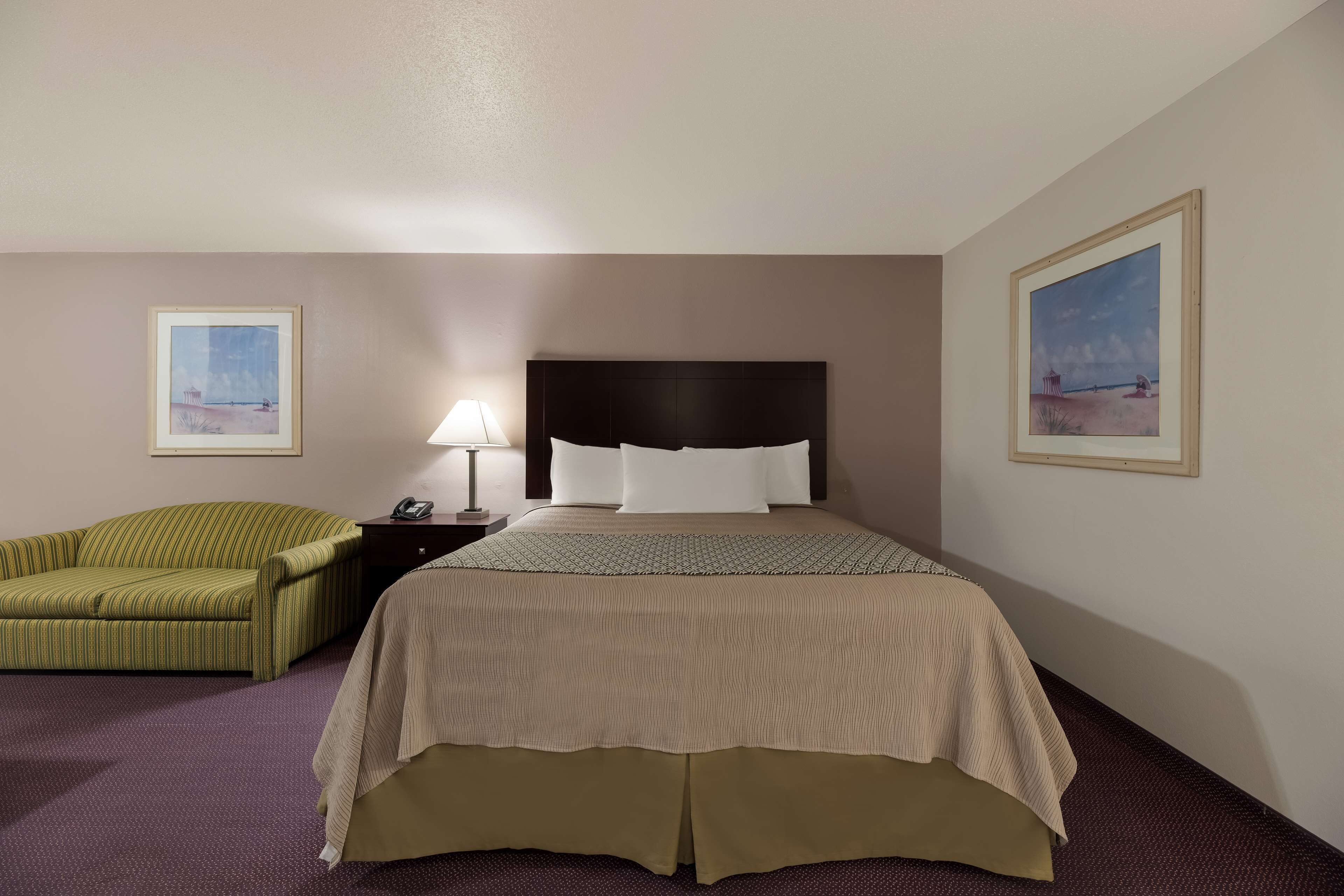 A hotel room with a large bed, a green striped sofa, a nightstand with a lamp, and two framed pictures on the walls.