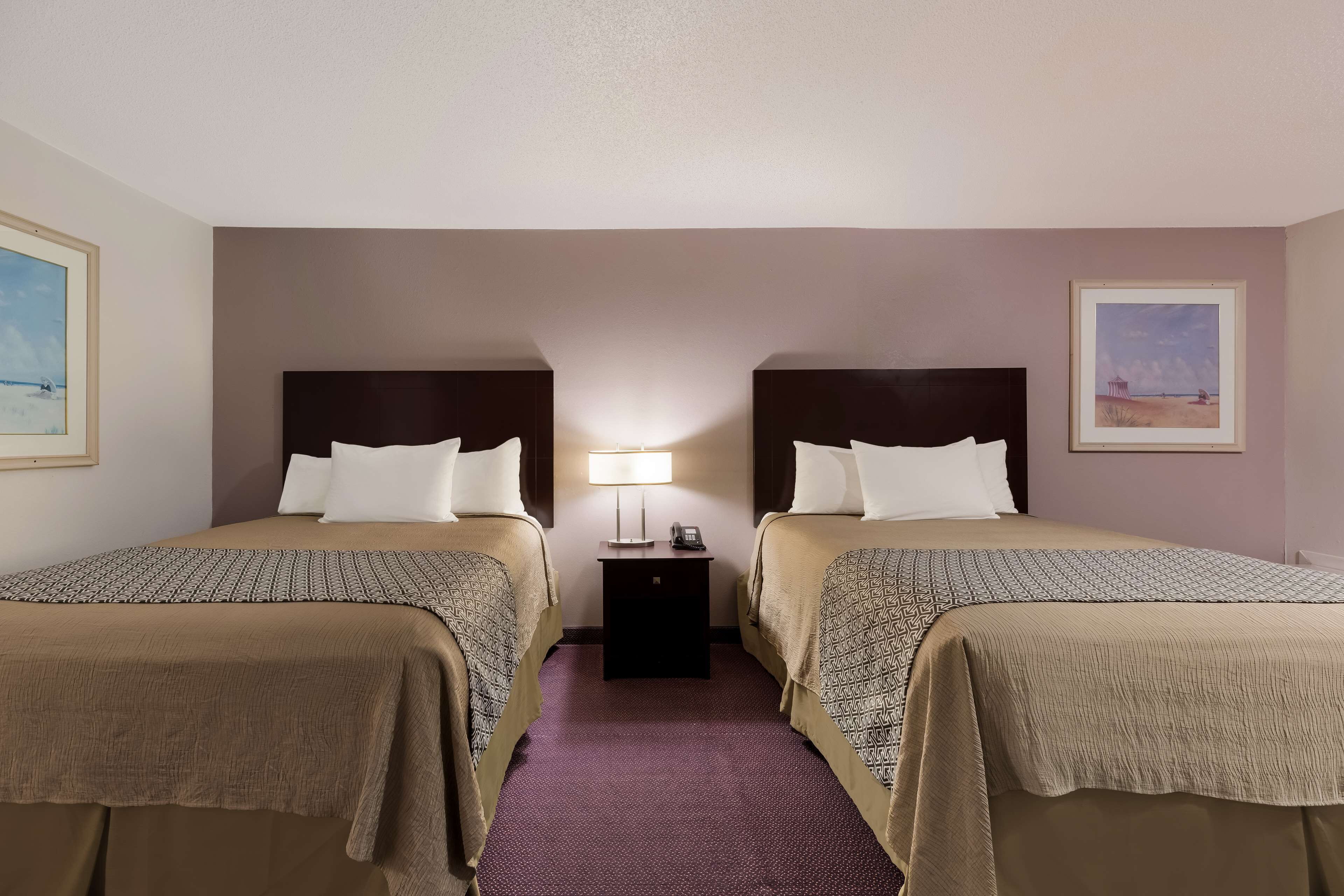 Habitación de hotel con dos camas, una mesita de noche con lámpara y cuadros en las paredes.