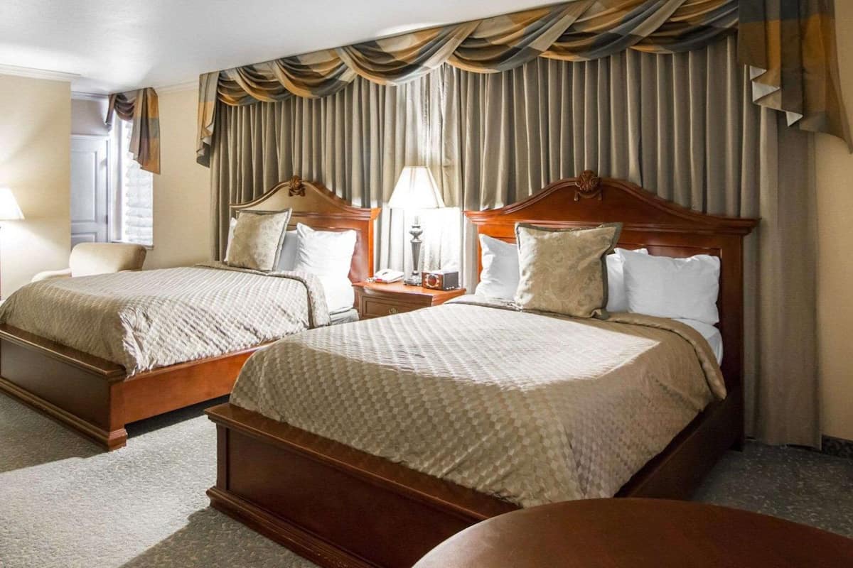 A hotel room with two double beds, wooden headboards, a nightstand with a lamp, and a large window with curtains.