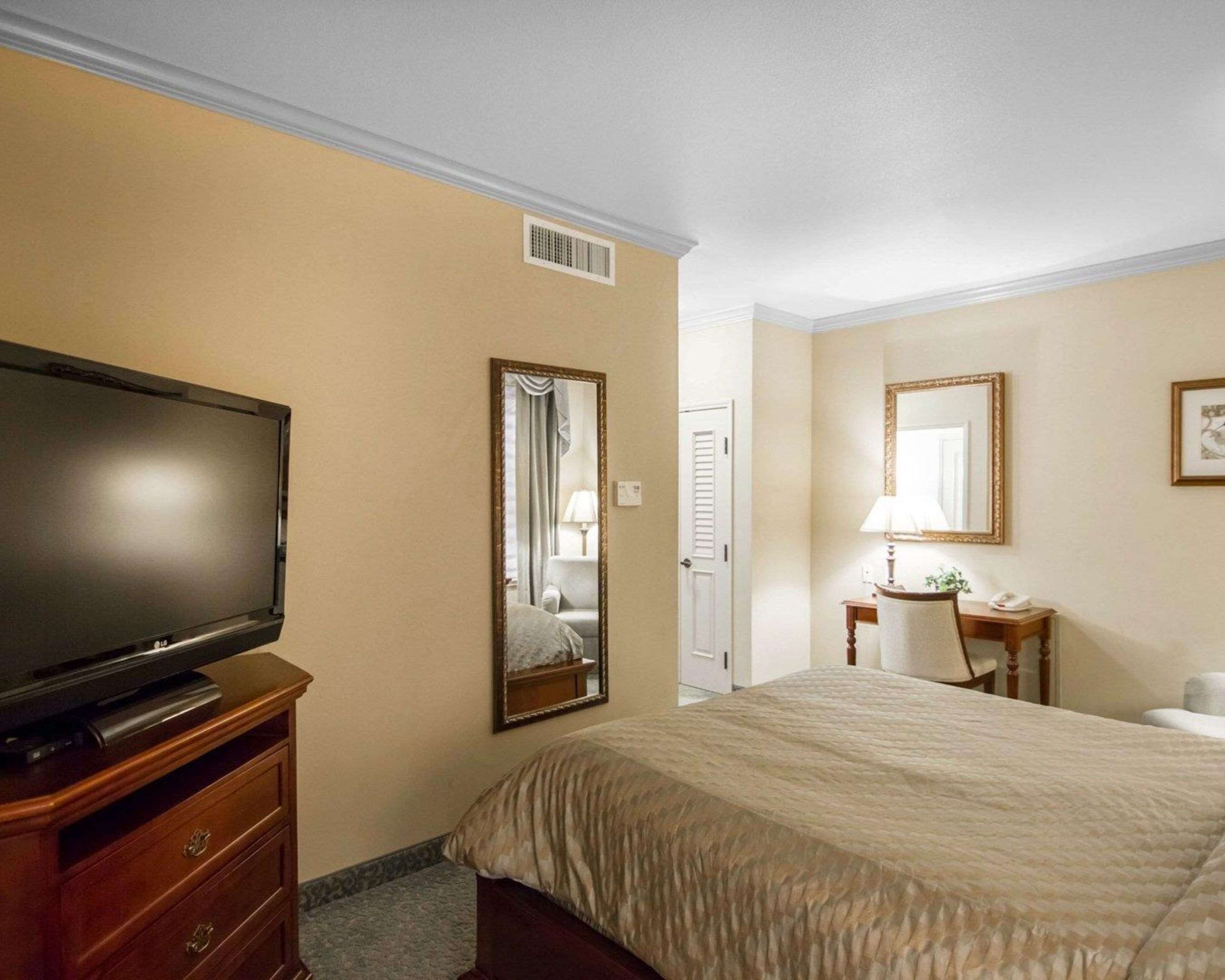 A hotel room with a bed, a television on a wooden dresser, a mirror, a desk with a lamp, and a chair.