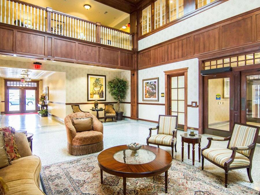 A spacious lobby with high ceilings, wooden paneling, and a central seating area with a round table. There are multiple chairs and a sofa, and a large window allows natural light to enter.