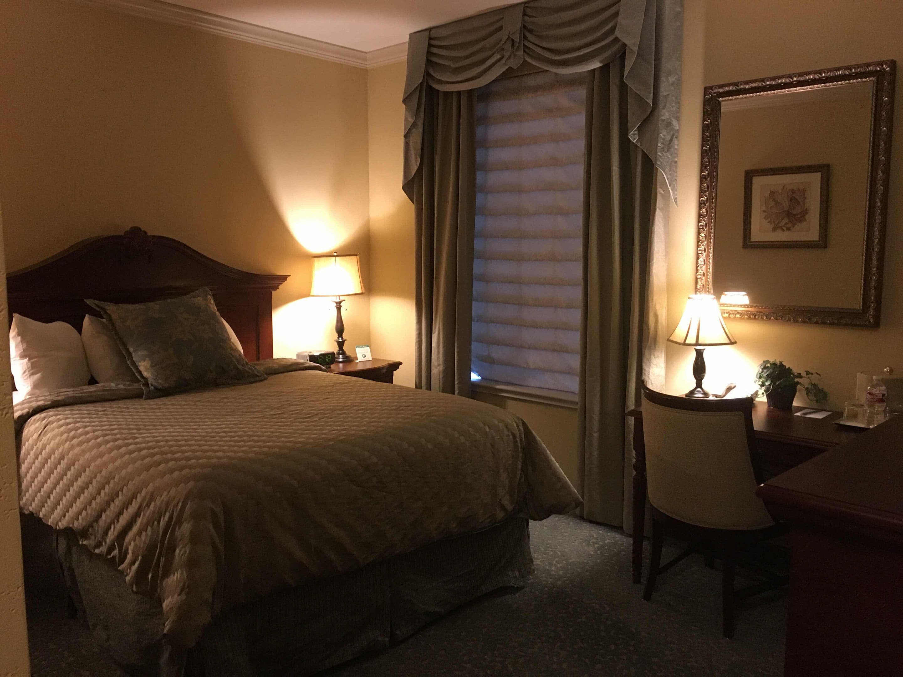A hotel room with a large bed, two bedside tables with lamps, a chair, a desk, a window with blinds, and a framed picture on the wall.