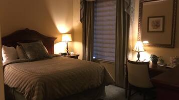 A hotel room with a large bed, two bedside tables with lamps, a chair, a desk, a window with blinds, and a framed picture on the wall.