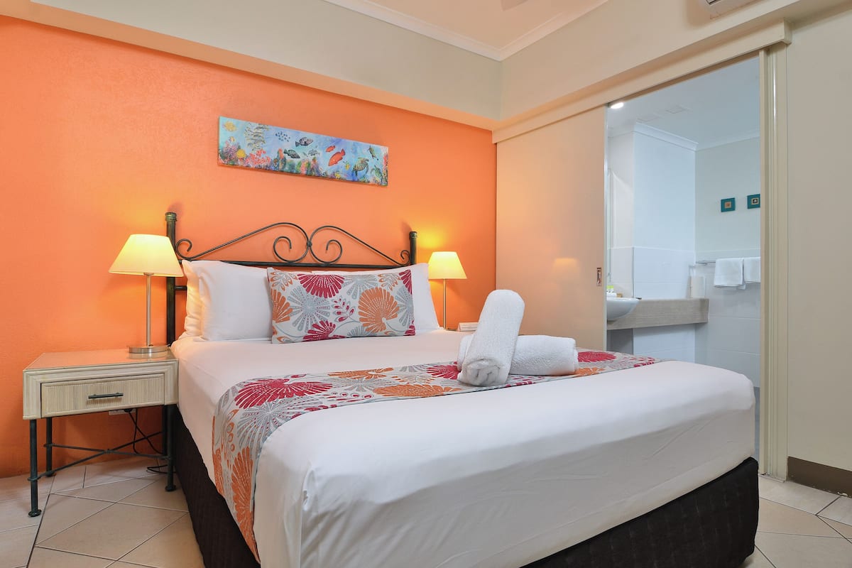 Habitación de hotel con cama, dos lámparas de noche, un cuadro en la pared y baño visible a través de una puerta de cristal.
