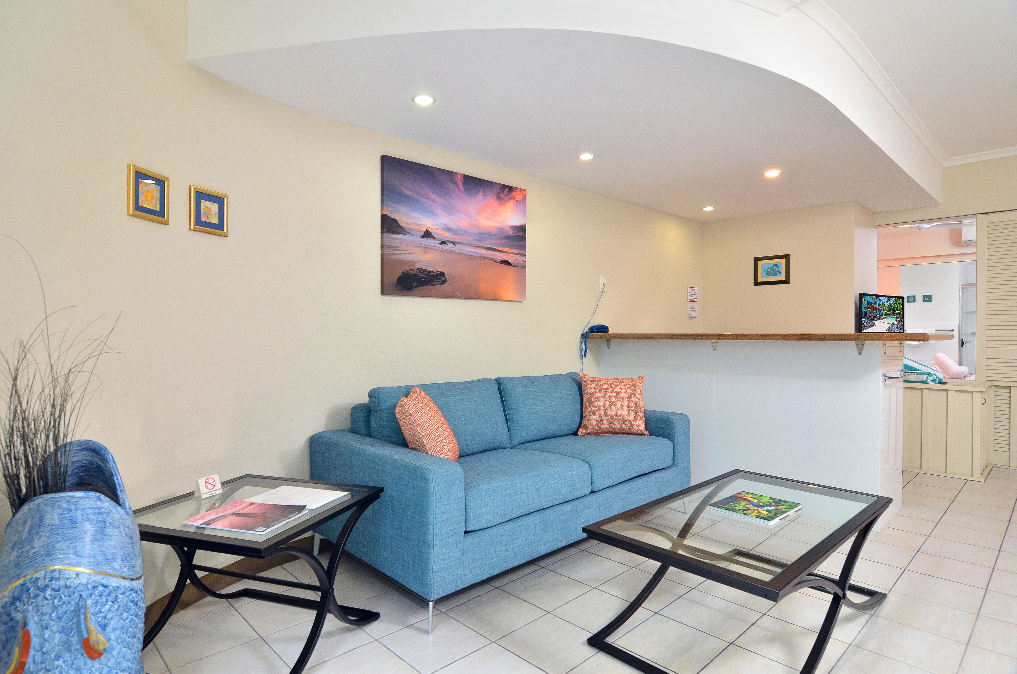 A living room with a blue sofa, glass coffee table, and a painting of a sunset.