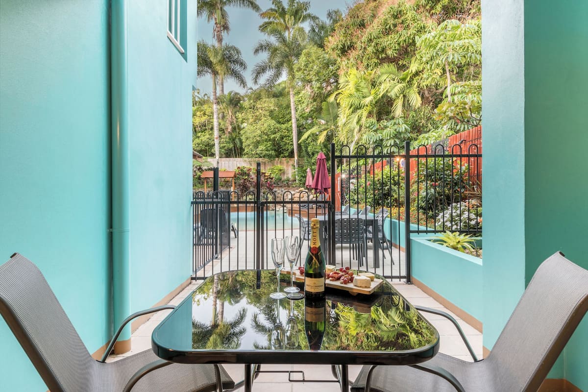Una mesa de vidrio con sillas, una botella de champán y una bandeja de frutas en un balcón con vista a una piscina y plantas tropicales.