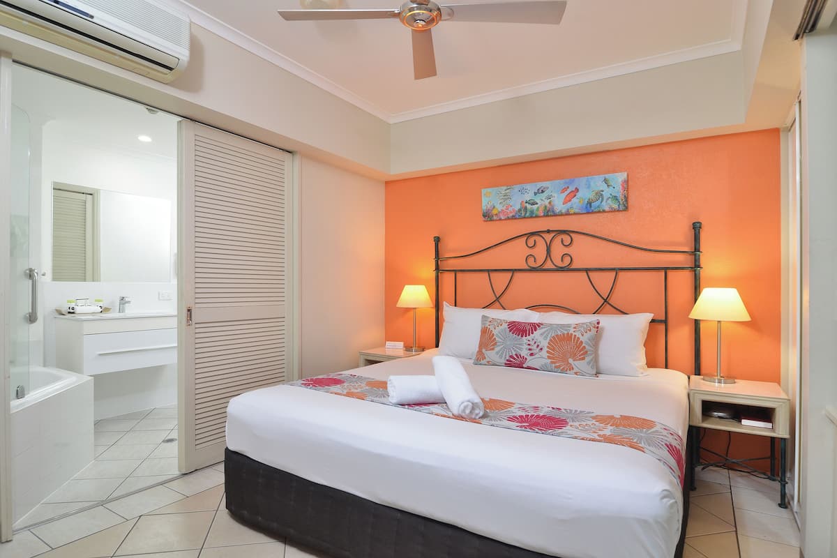 Una habitación de hotel con una cama, dos lámparas de noche, un ventilador de techo, un equipo de aire acondicionado, un baño con bañera y ducha.
