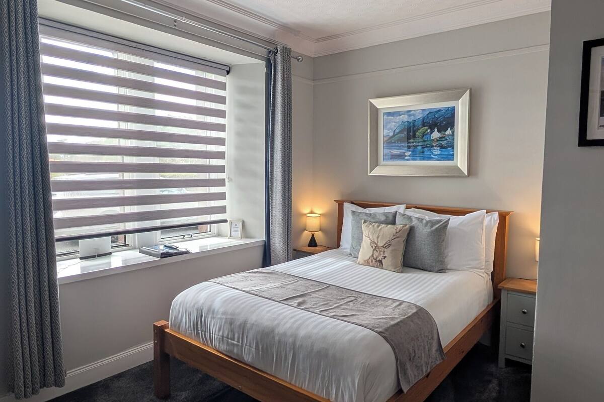 A bedroom with a large bed, a window with blinds, a painting on the wall, and a ceiling with a decorative pattern.
