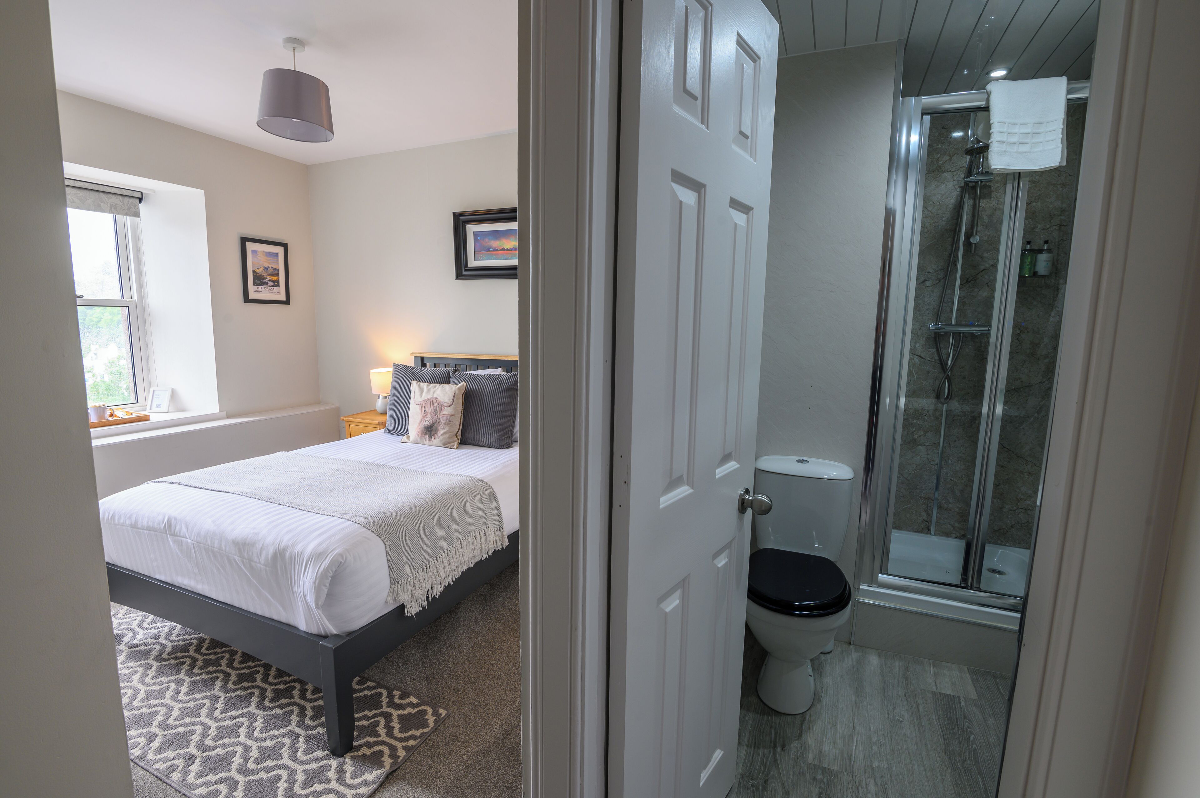Un dormitorio con cama, mesita de noche, ventana y puerta que da a un baño con ducha e inodoro.