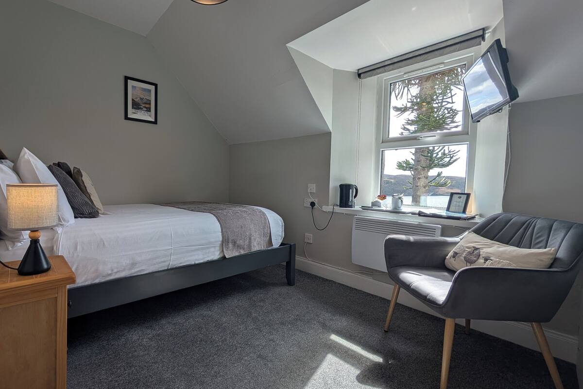 A bedroom with a bed, a desk, a chair, a TV, and a window with a view.