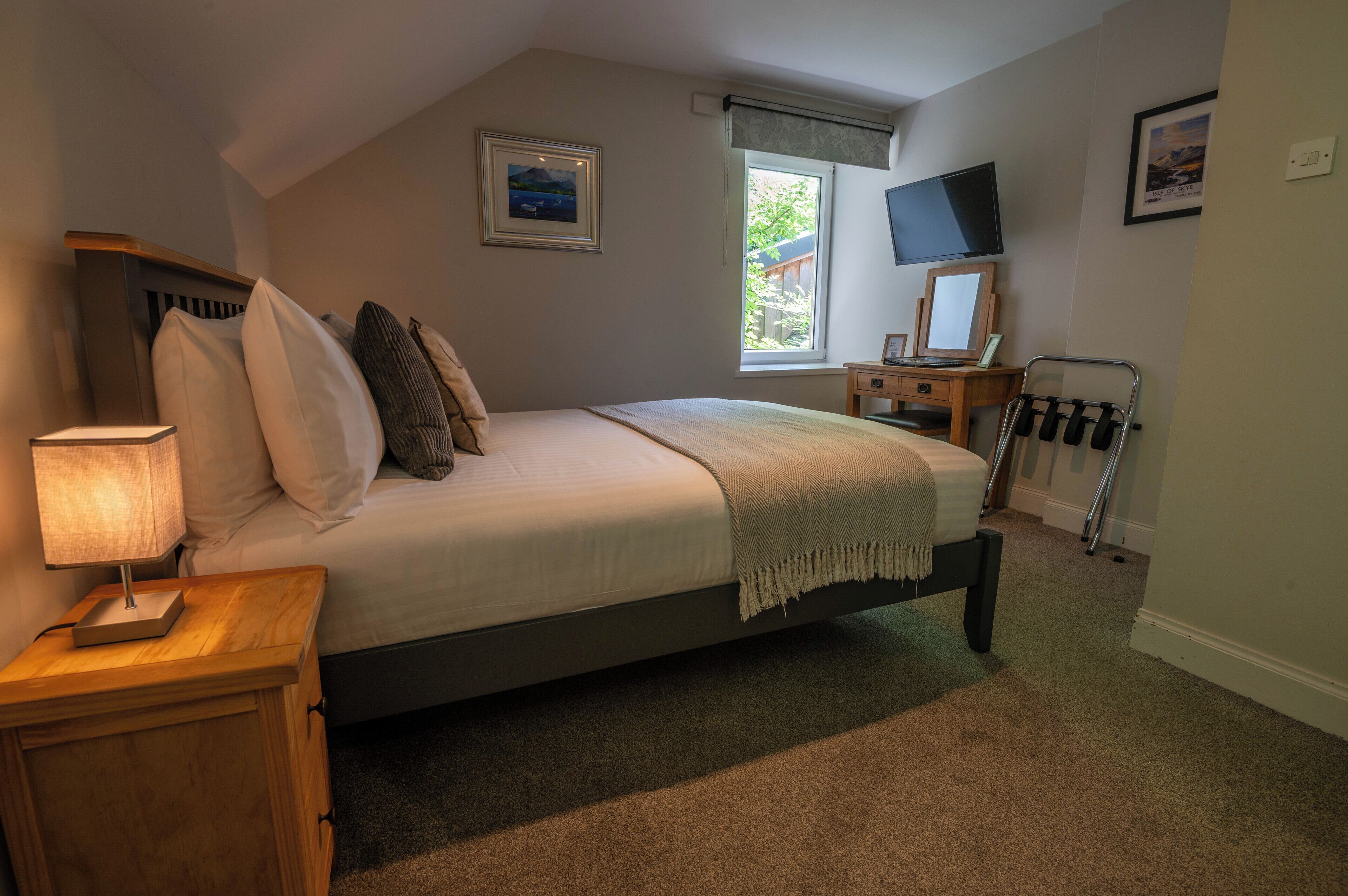 A bedroom with a bed, bedside table, lamp, TV, and a window with a view of greenery.