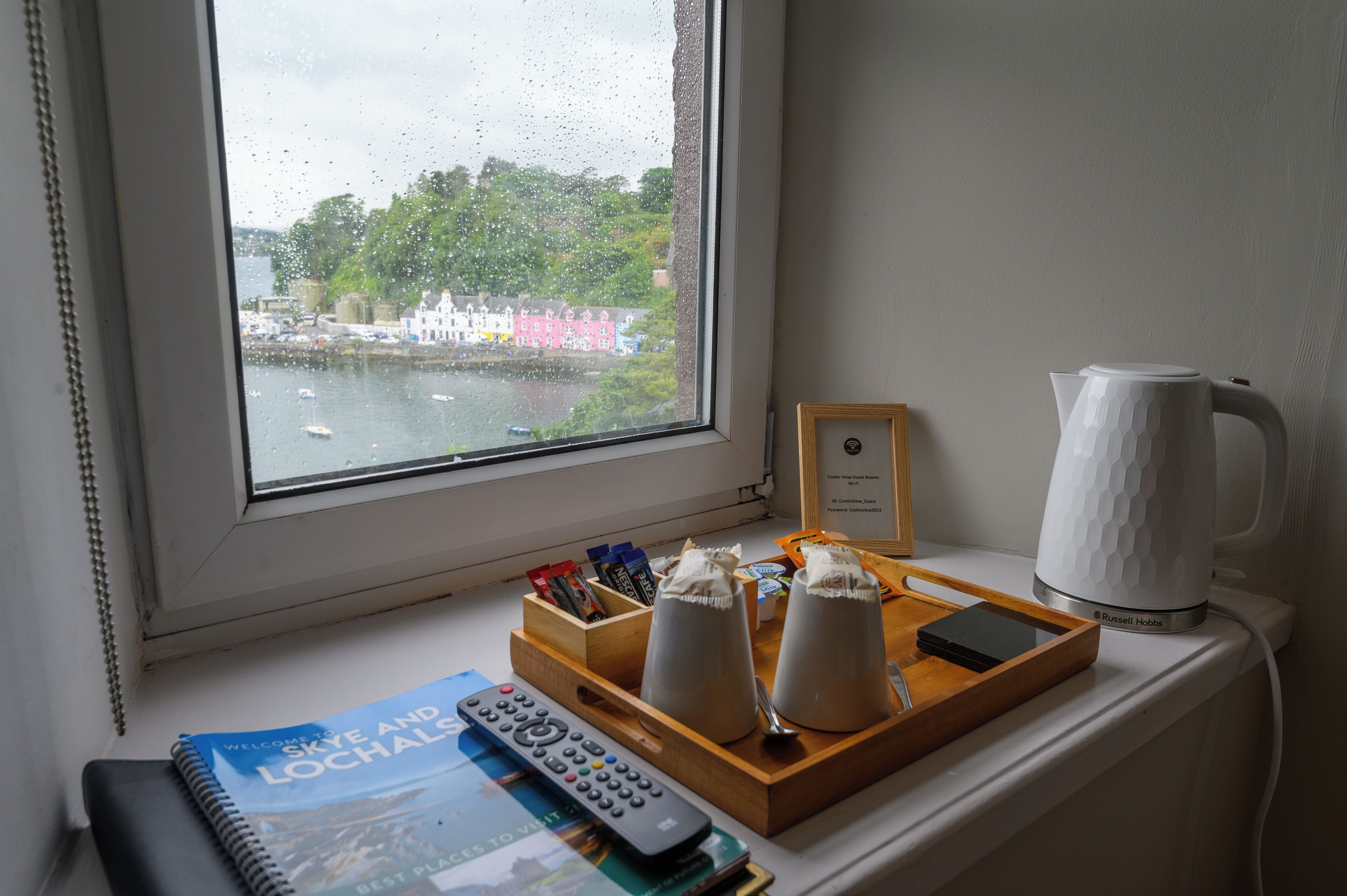 A window with a view of a body of water and buildings, a tray with cups and a remote control, and a kettle on a radiator.