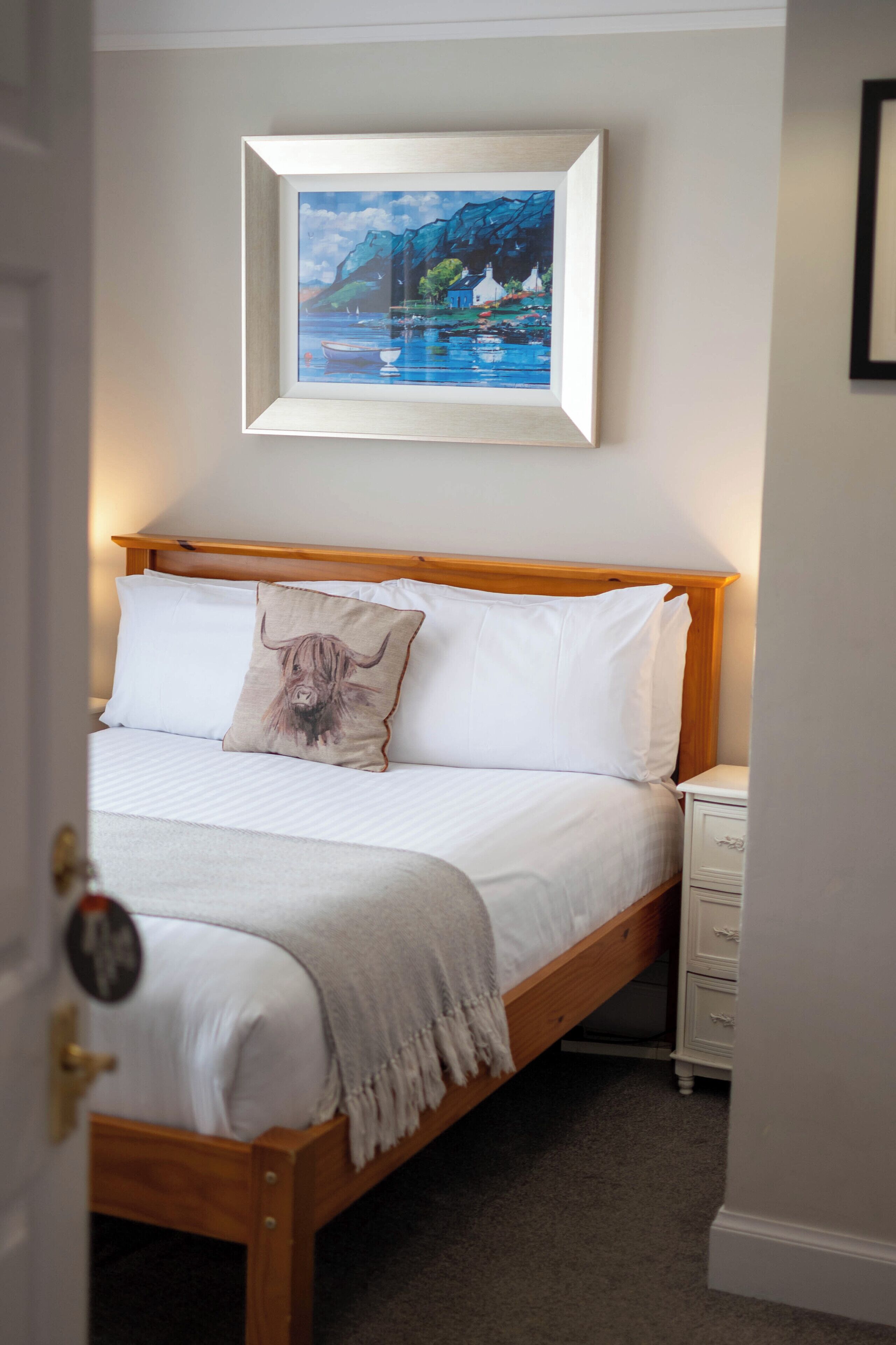 Un letto rifatto con cura, con una testiera in legno, un quadro appeso sopra e un comodino con un cassetto.