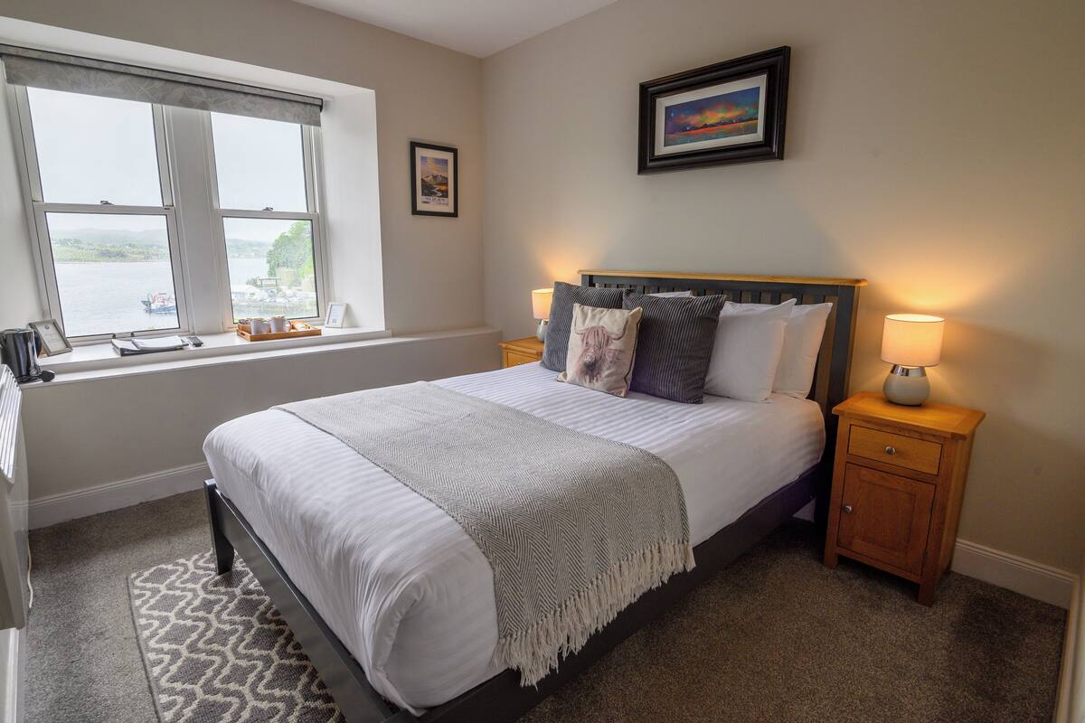A neatly made bed with a grey and white striped blanket, a wooden nightstand with a lamp, and a window with a view of a body of water.